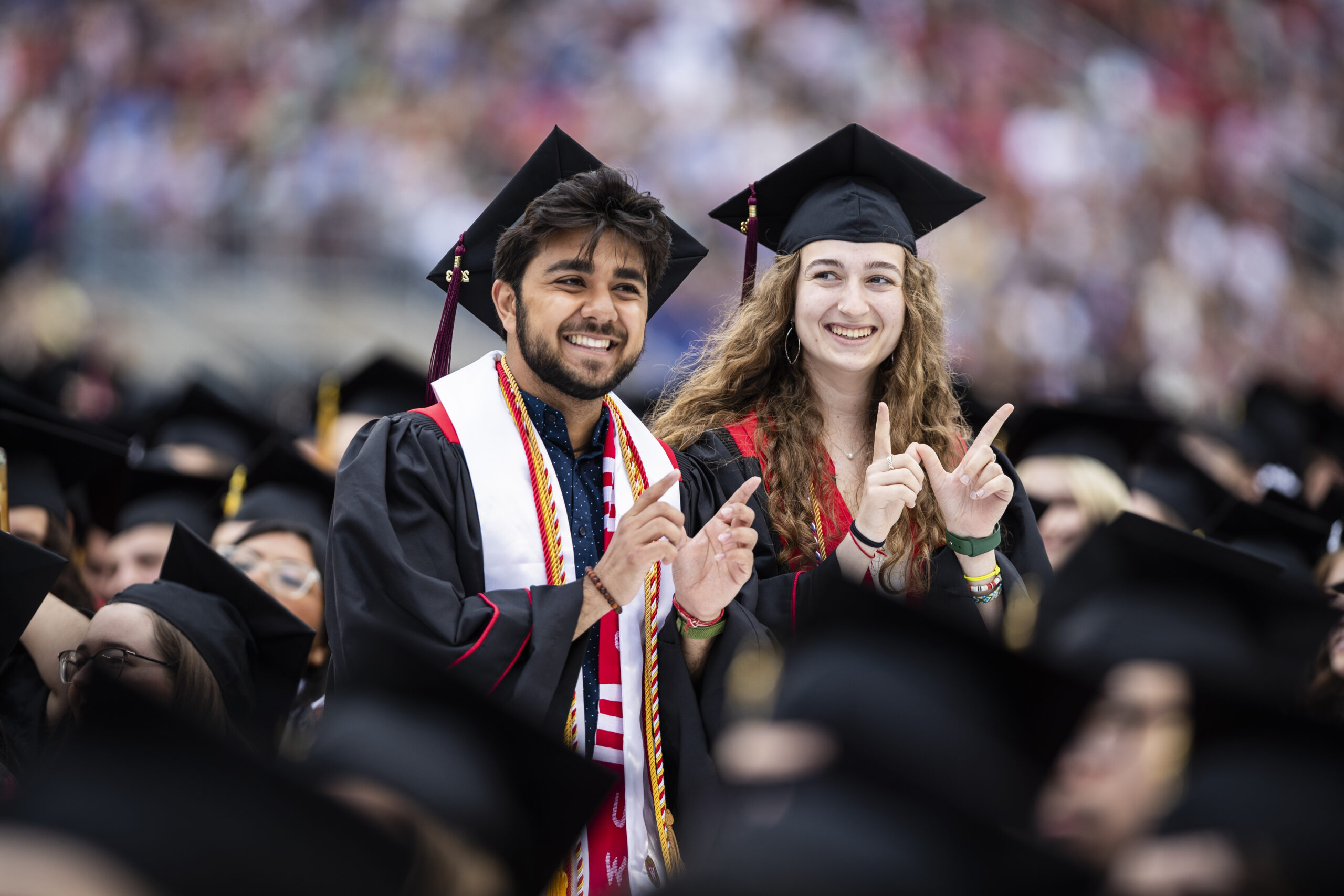 Two graduates making a Wisconsin "W" with their hands, dressed in their cap & gown at commencement